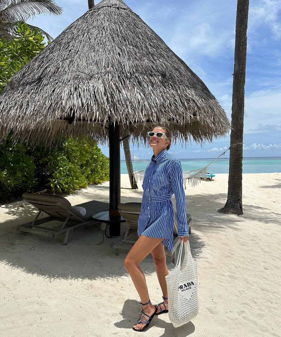 Pessoa na praia com vestido listrado azul e sandálias tipo gladiadora, segura bolsa clara ao lado de cabana de palha.