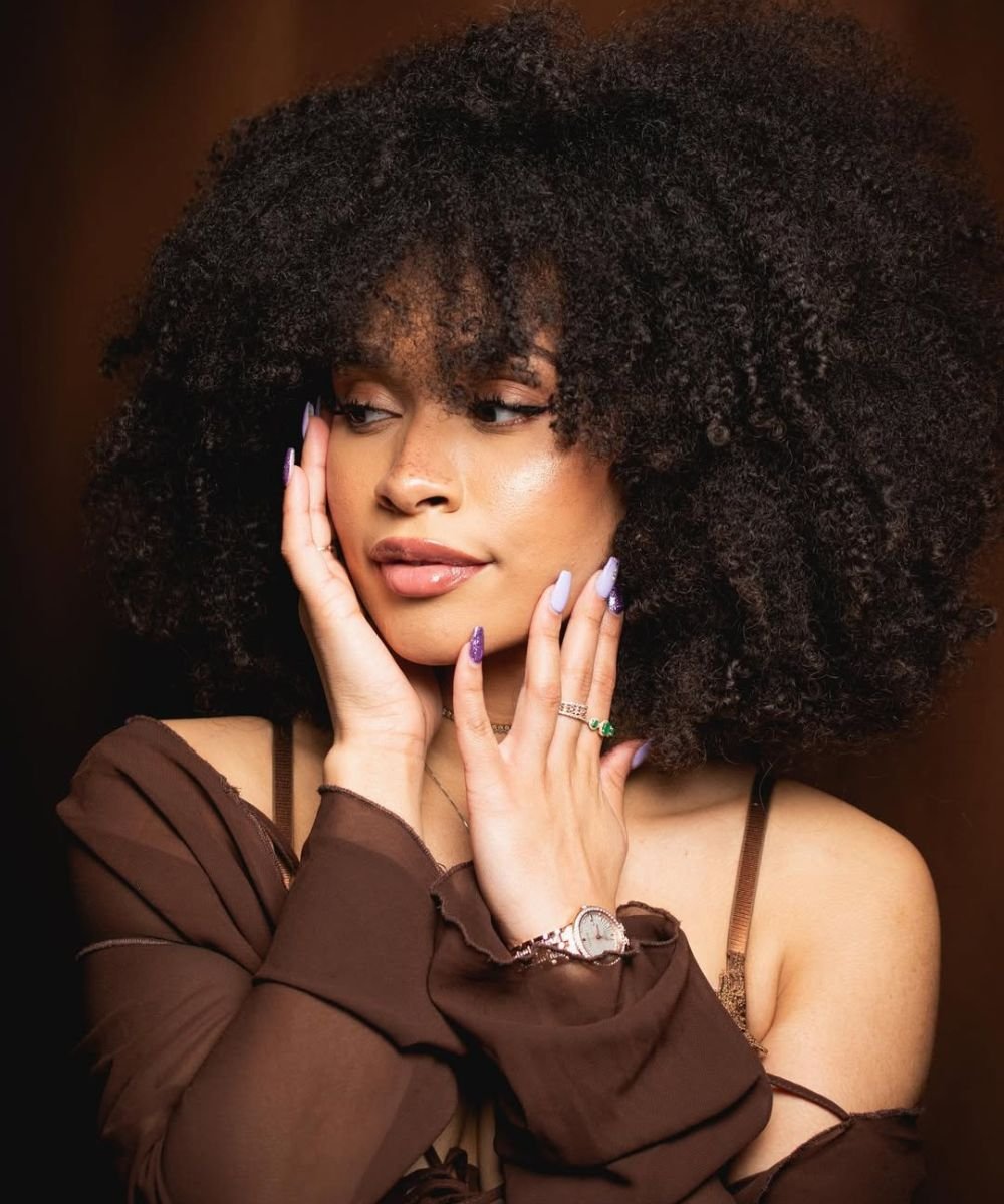 Mulher com cabelo volumoso, bem hidratado, com mãos próximas ao rosto e unhas pintadas depois da hidratação para cabelo crespo.