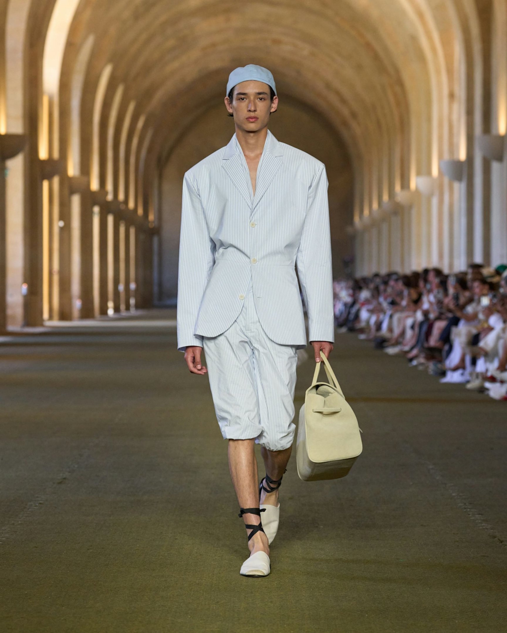 Desfile de moda com modelo usando terno listrado e shorts, segurando mala bege. Ambiente iluminado e elegante.