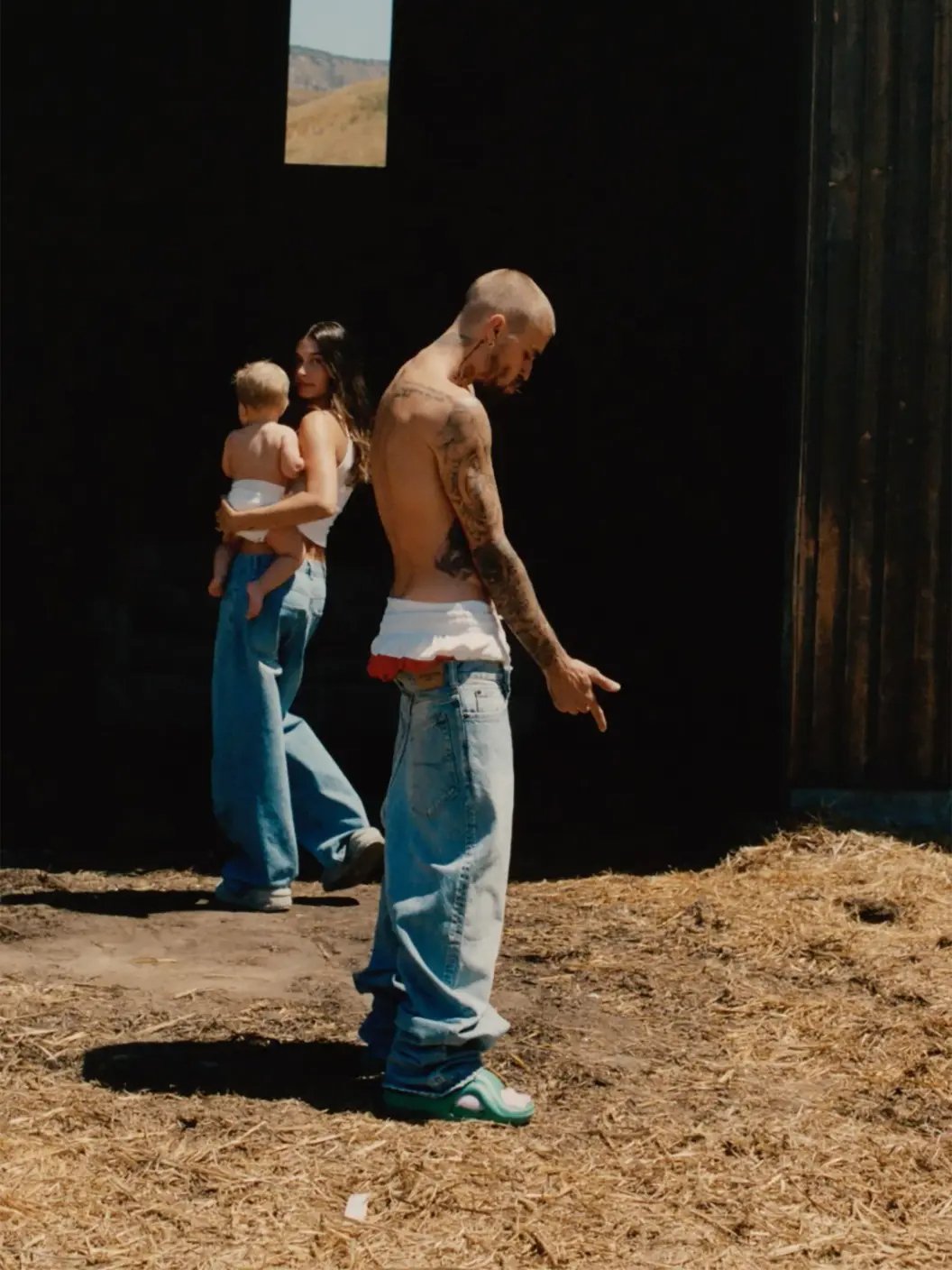 Justin Bieber sem camisa e calça jeans olhando para o chão; mulher e criança ao fundo.