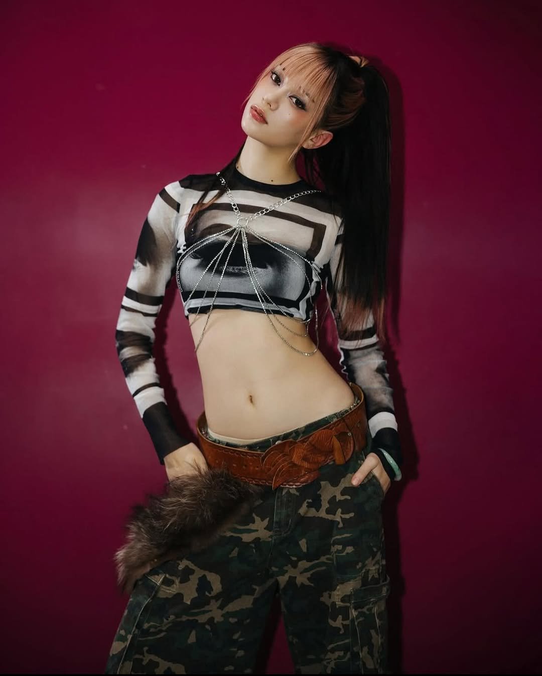 Ela usa cropped preto e calça camuflada, posando contra parede roxa. katseye