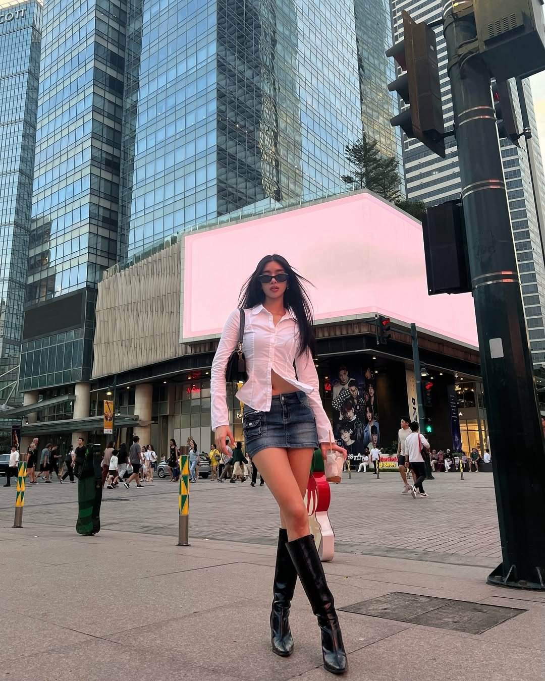 Pessoa de óculos escuros e roupa estilosa em frente a arranha-céus, com outdoor ao fundo. Katseye