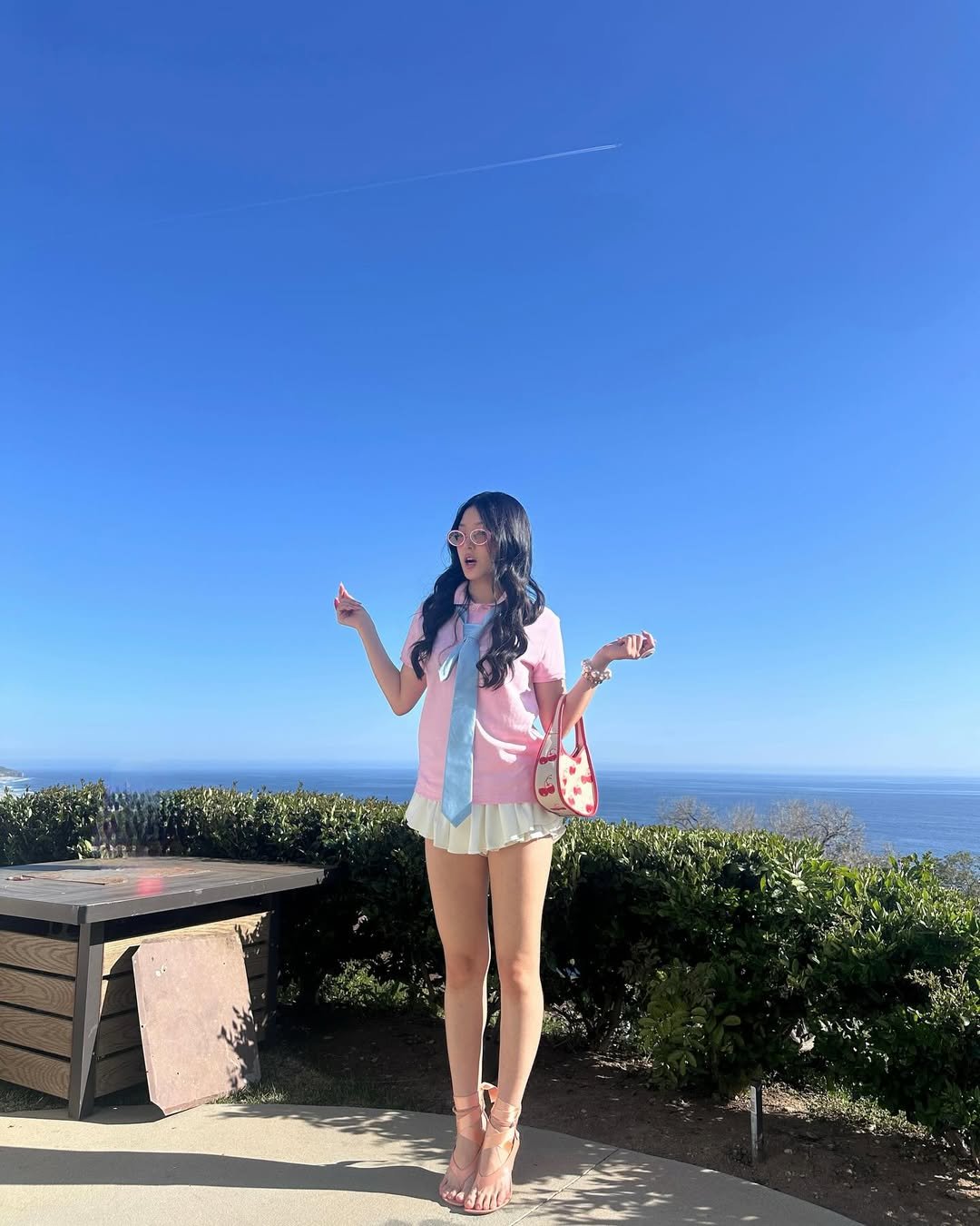 Pessoa com saia clara e blusa rosa, em um jardim com vista para o mar. Ideal para passeios ensolarados e katseye.