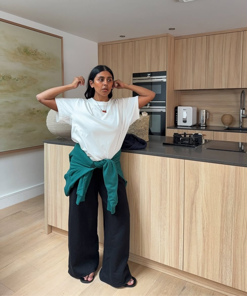 Mulher em cozinha vestindo camiseta branca e calça preta, casaco verde amarrado na cintura. Estilo descontraído.