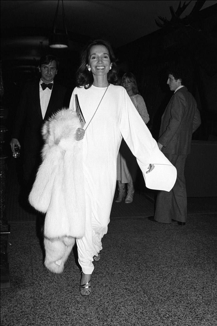 Lee Radziwillusando vestido branco e casaco de pele, cercada por outras pessoas em um evento social.