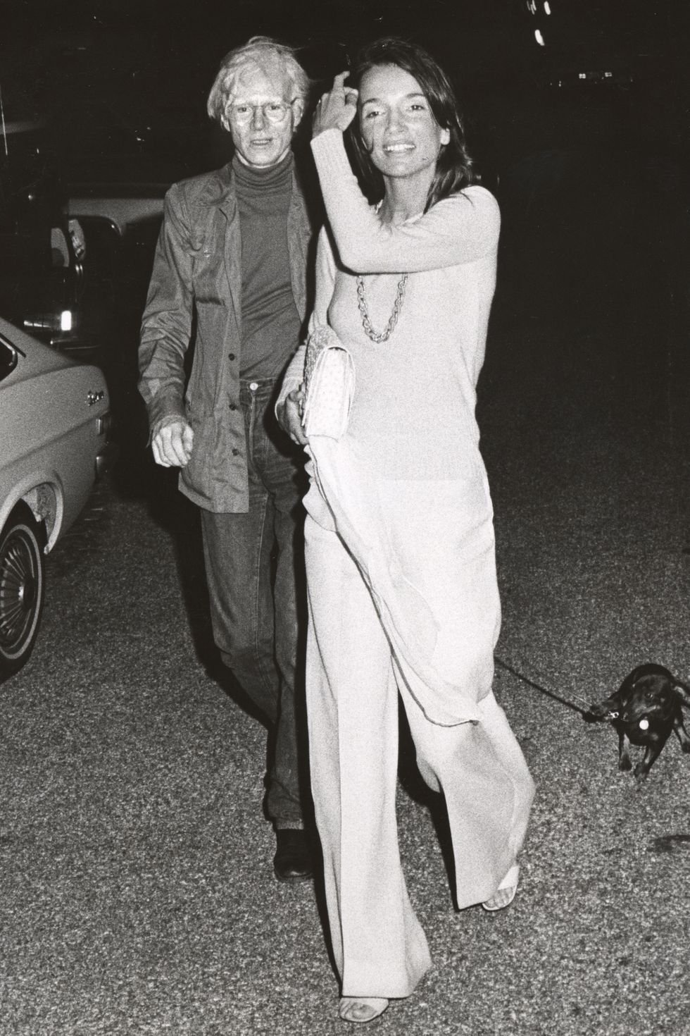 Lee Radziwill sorrindo caminha com homem, segurando uma bolsa e a trela de um cachorro.