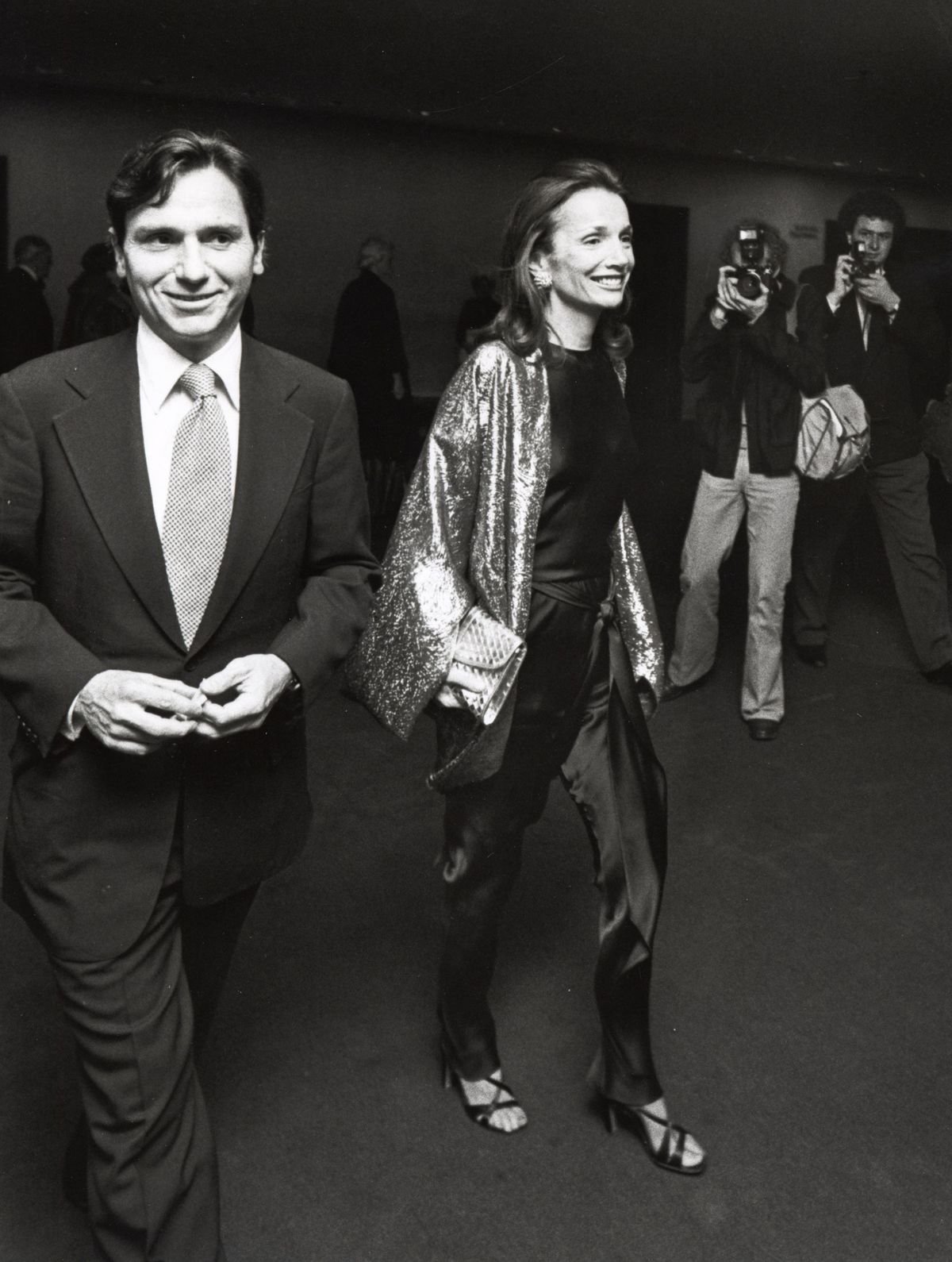 Lee Radziwill. Duas pessoas elegantes caminham sorrindo enquanto fotógrafos capturam o momento em um evento social.