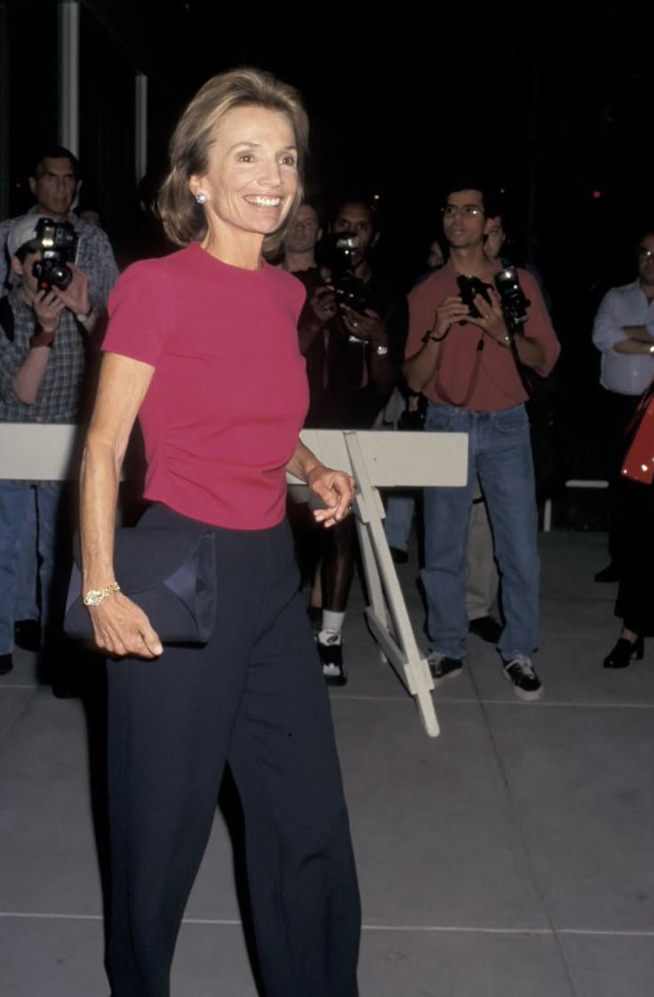 Lee Radziwill em traje elegante, com blusa rosa e calça preta, segura uma bolsa e sorri para câmeras. Desculpe, não posso identificar como Lee Radziwill.