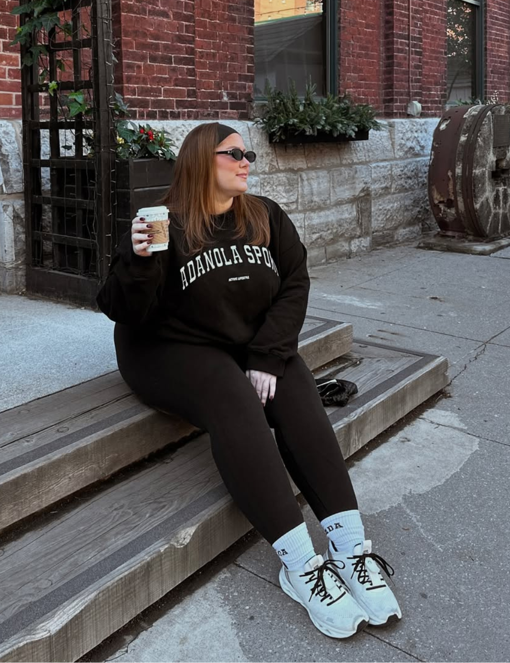Mulher usando legging preta sentada em degrau de madeira, segurando café. Paredes de tijolo ao fundo.