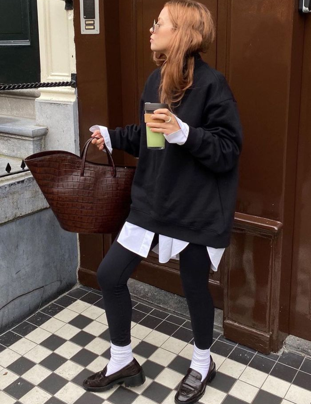 Mulher com legging preta, moletom escuro, segurando bebida e bolsa marrom, em calçada quadriculada.
