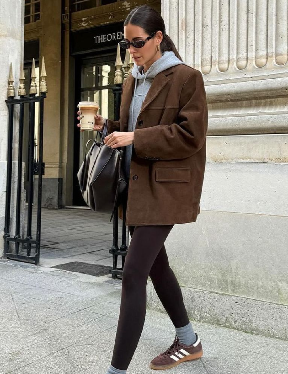 Pessoa usando legging preta, blazer marrom e tênis, segurando bebida. Edifício clássico ao fundo.