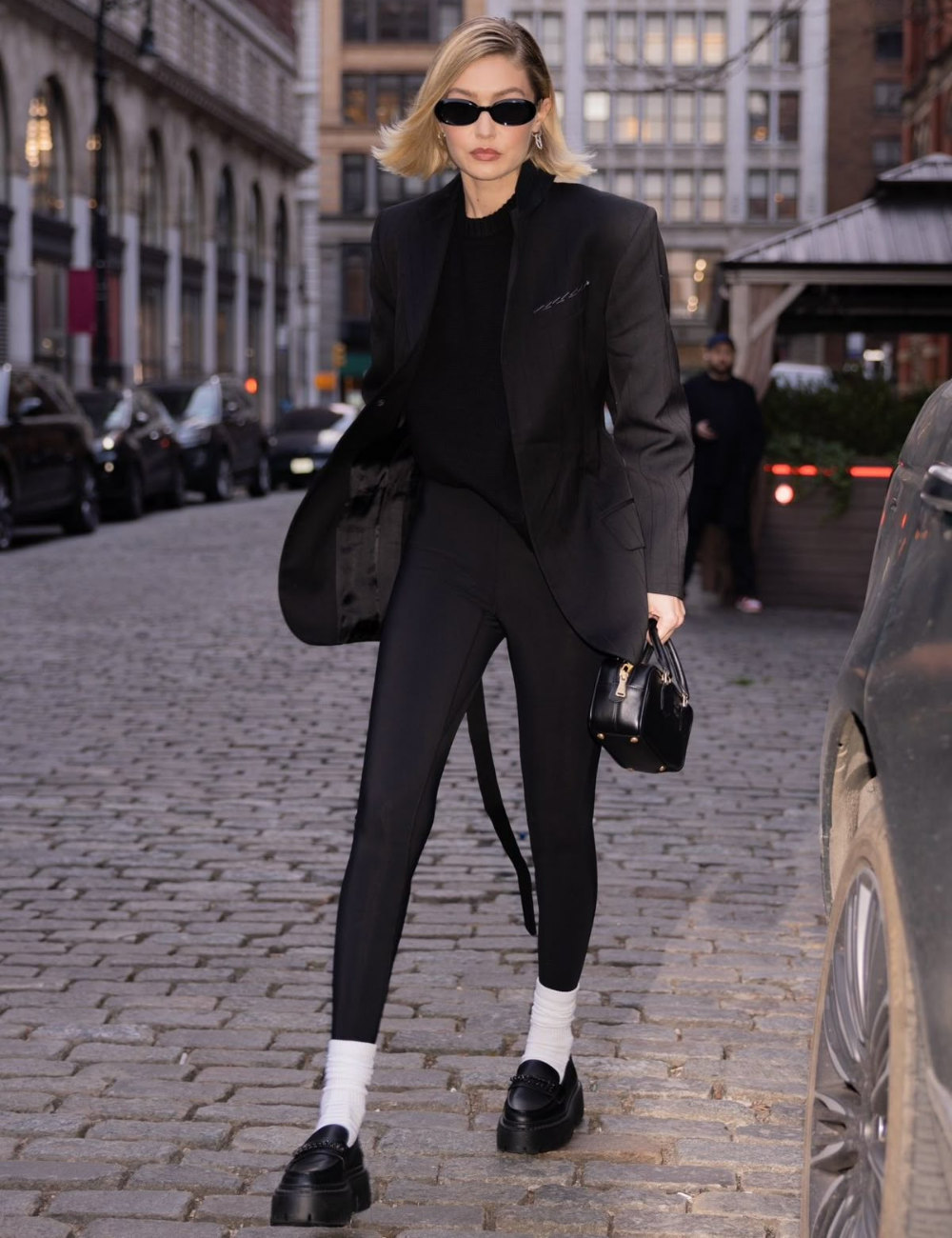 Pessoa usando blazer preto, legging e sapatos escuros em rua com paralelepípedos. Ela segura uma bolsa pequena.