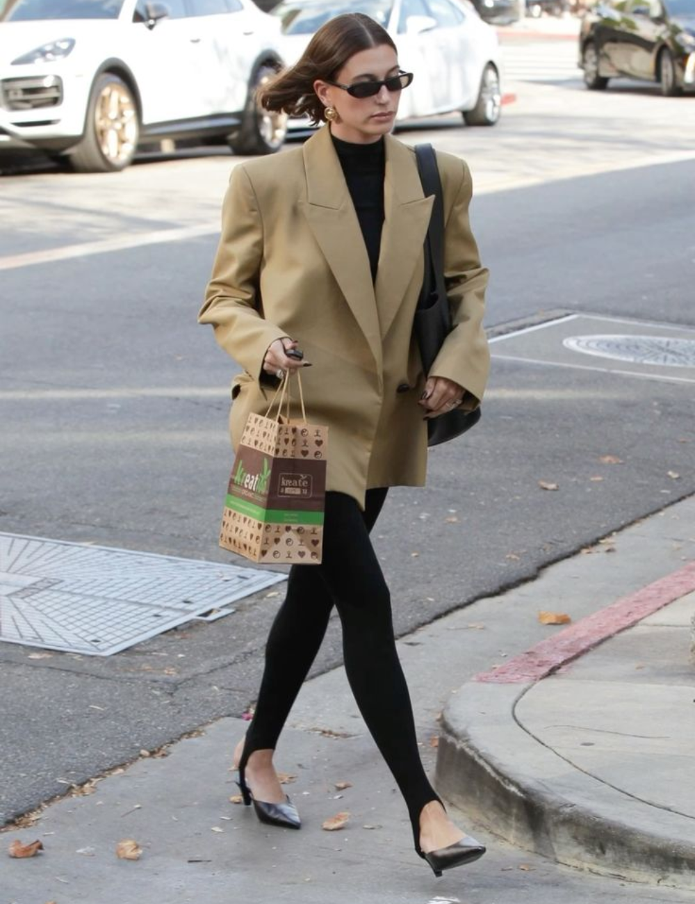 Mulher usando legging preta, blazer bege e óculos escuros, caminhando na calçada e carregando sacola.