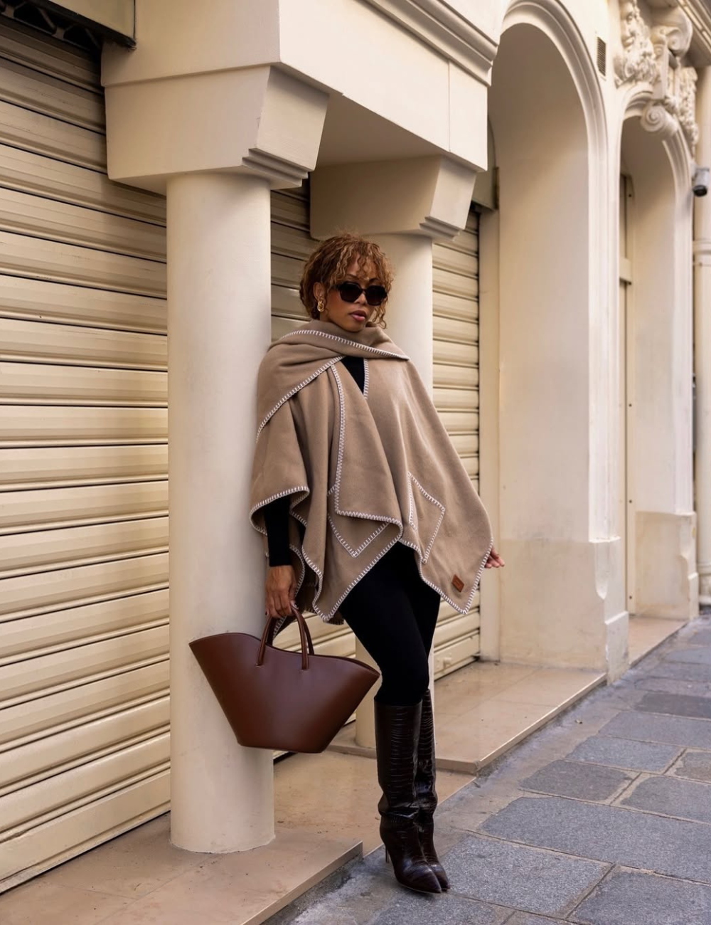 Pessoa com poncho bege e legging preta, segurando bolsa marrom, encostada em coluna em área urbana.