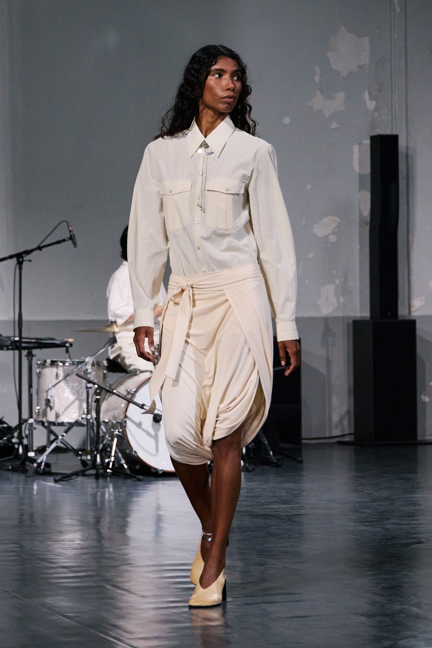Modelo vestindo calça balonê bege com camisa branca em desfile de moda. Bateria ao fundo.