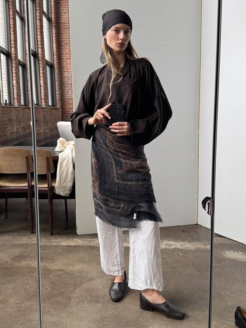 Pessoa em frente ao espelho vestindo roupas largas e escuras, com lenço preto na cabeça e calças brancas.