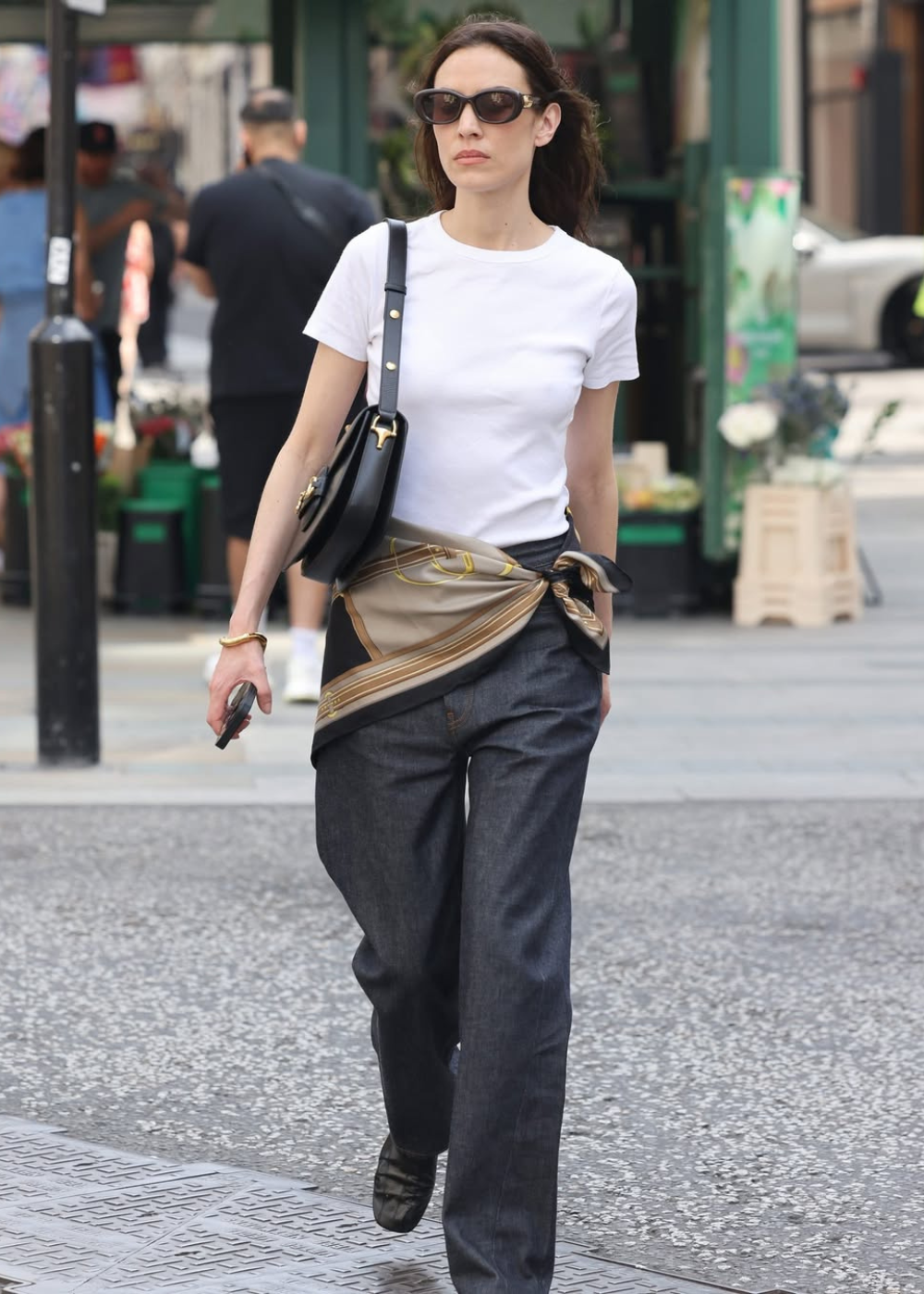 Mulher usando camiseta branca e óculos escuros, com lenço no quadril sobre calça escura, andando pela rua.