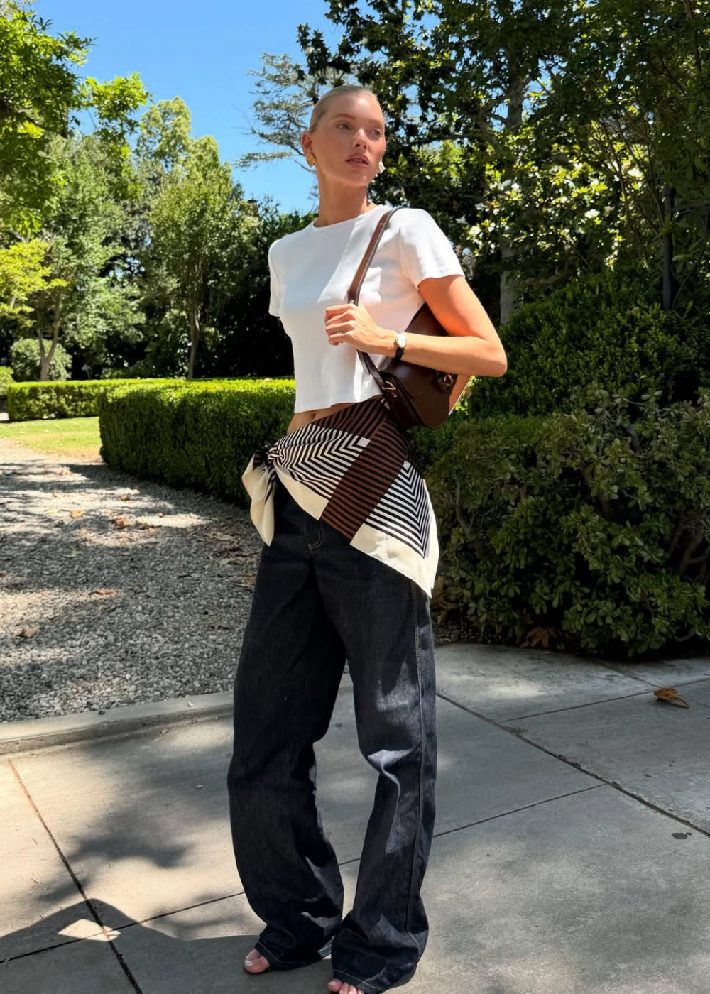 Mulher em pé com lenço amarrado no quadril, usando blusa branca e calça larga, em ambiente ao ar livre com árvores.