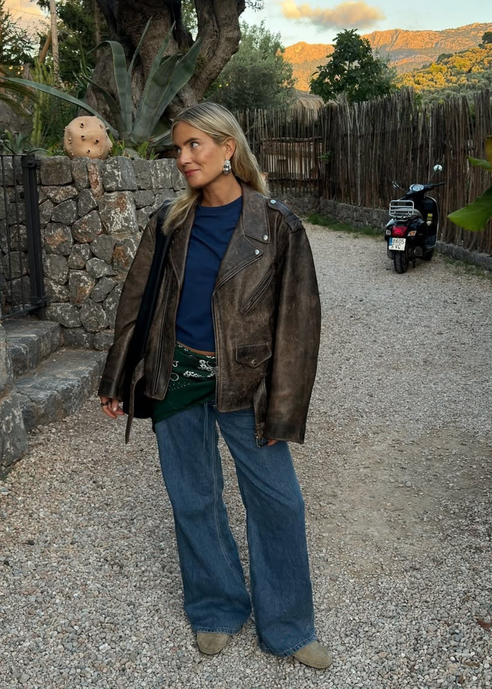 Mulher de jaqueta de couro, blusa azul e lenço verde no quadril, em local externo com moto ao fundo.