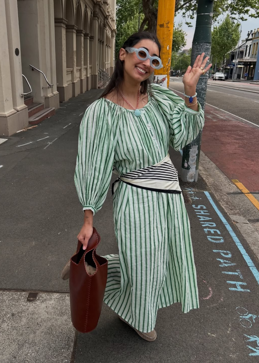 Mulher sorridente com óculos brancos, vestido listrado verde e lenço no quadril, acenando na rua.