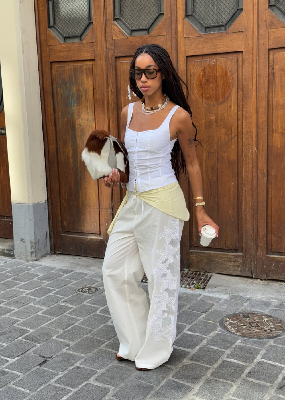 Jovem com lenço amarrado no quadril, segurando um café, em frente a porta de madeira.