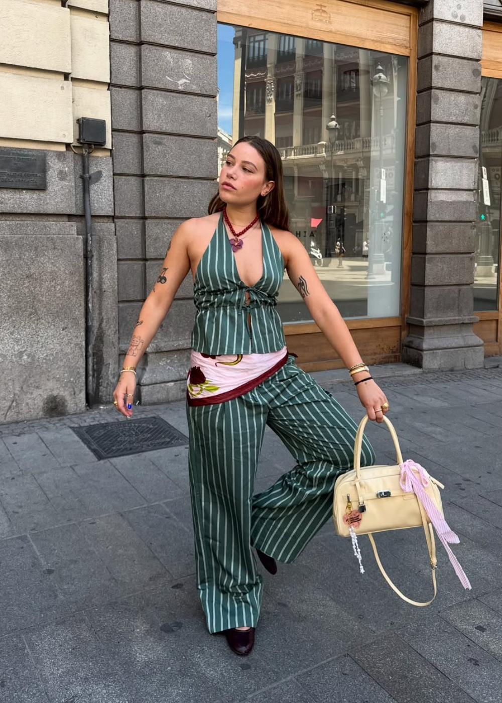 Pessoa com roupa verde listrada e lenço rosa no quadril, segurando uma bolsa bege enquanto caminha na calçada.
