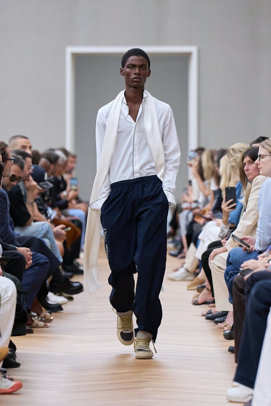 Modelo desfila usando camisa branca e calça azul, com um lenço leve ao redor do pescoço. Estudos sentados ao redor.