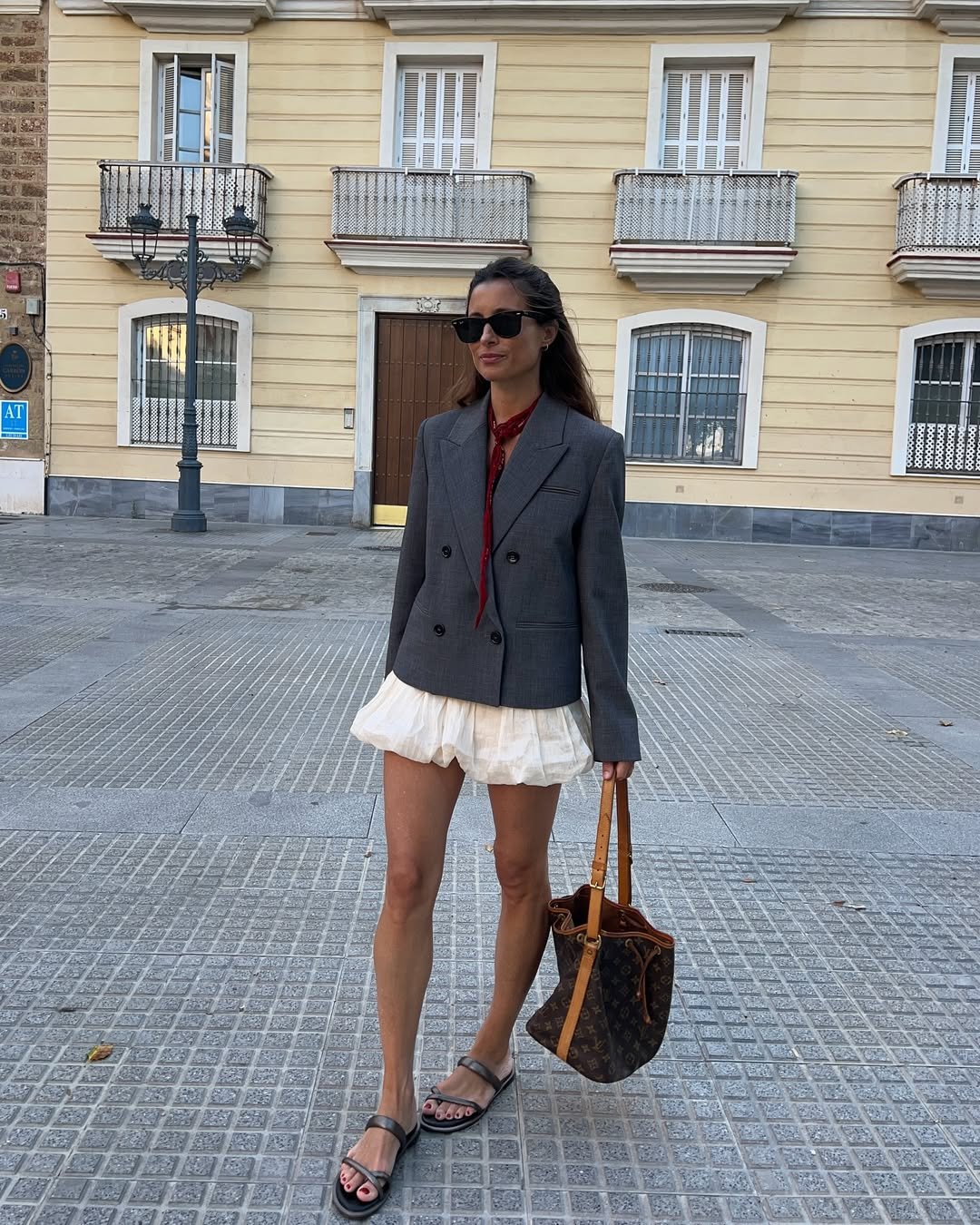 Pessoa de óculos escuros e lenço vermelho usa blazer cinza e saia branca em frente a prédio com fachadas clássicas.