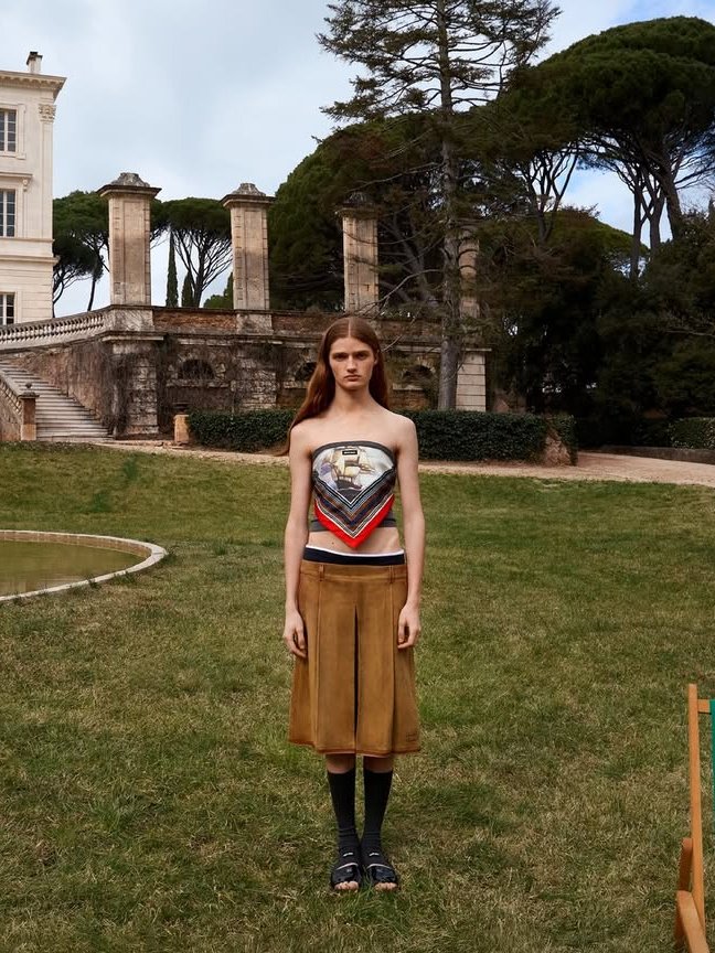 Jovem em jardim veste blusa estilo lenço, saia marrom, meias pretas e sandálias, com uma mansão ao fundo.