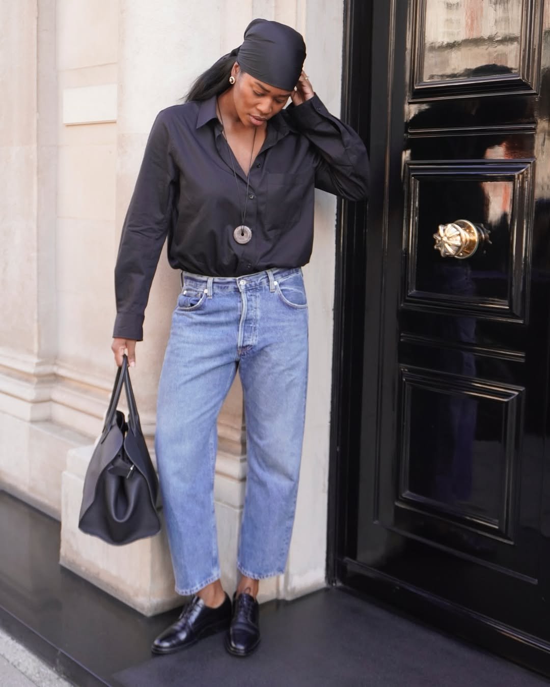 Mulher com camisa preta, jeans e lenço na cabeça segura bolsa ao lado de porta preta.