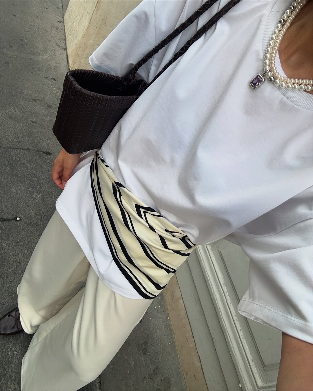Blusa branca com colar de pérolas e lenço listrado na cintura, combinados com bolsa escura e calça clara.