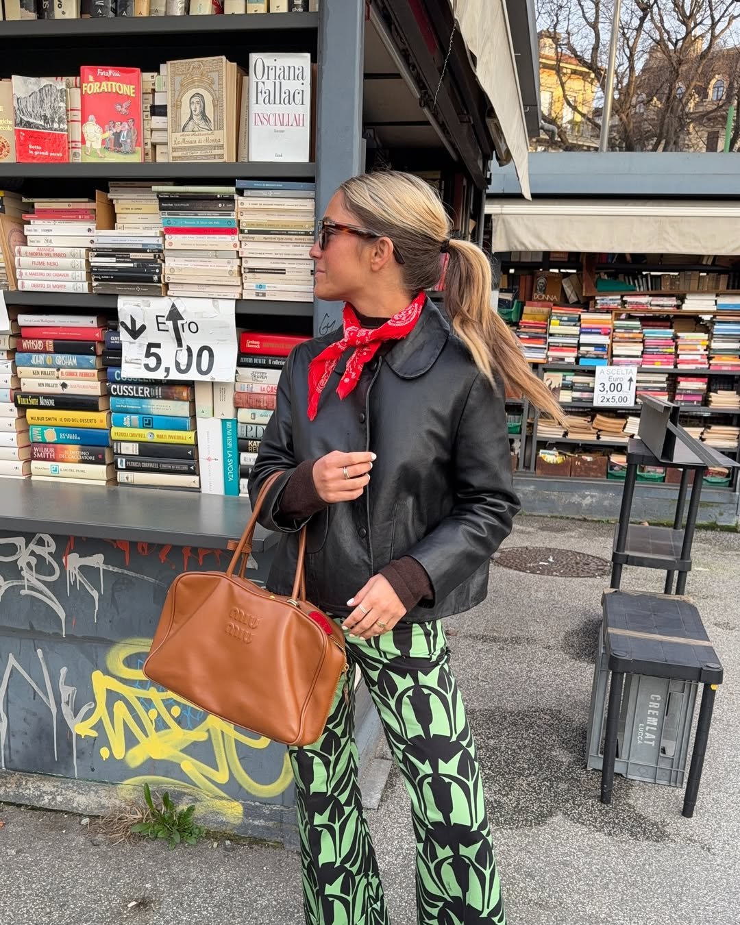 Mulher com lenço vermelho no pescoço está em frente a barraca de livros, segurando uma bolsa marrom.