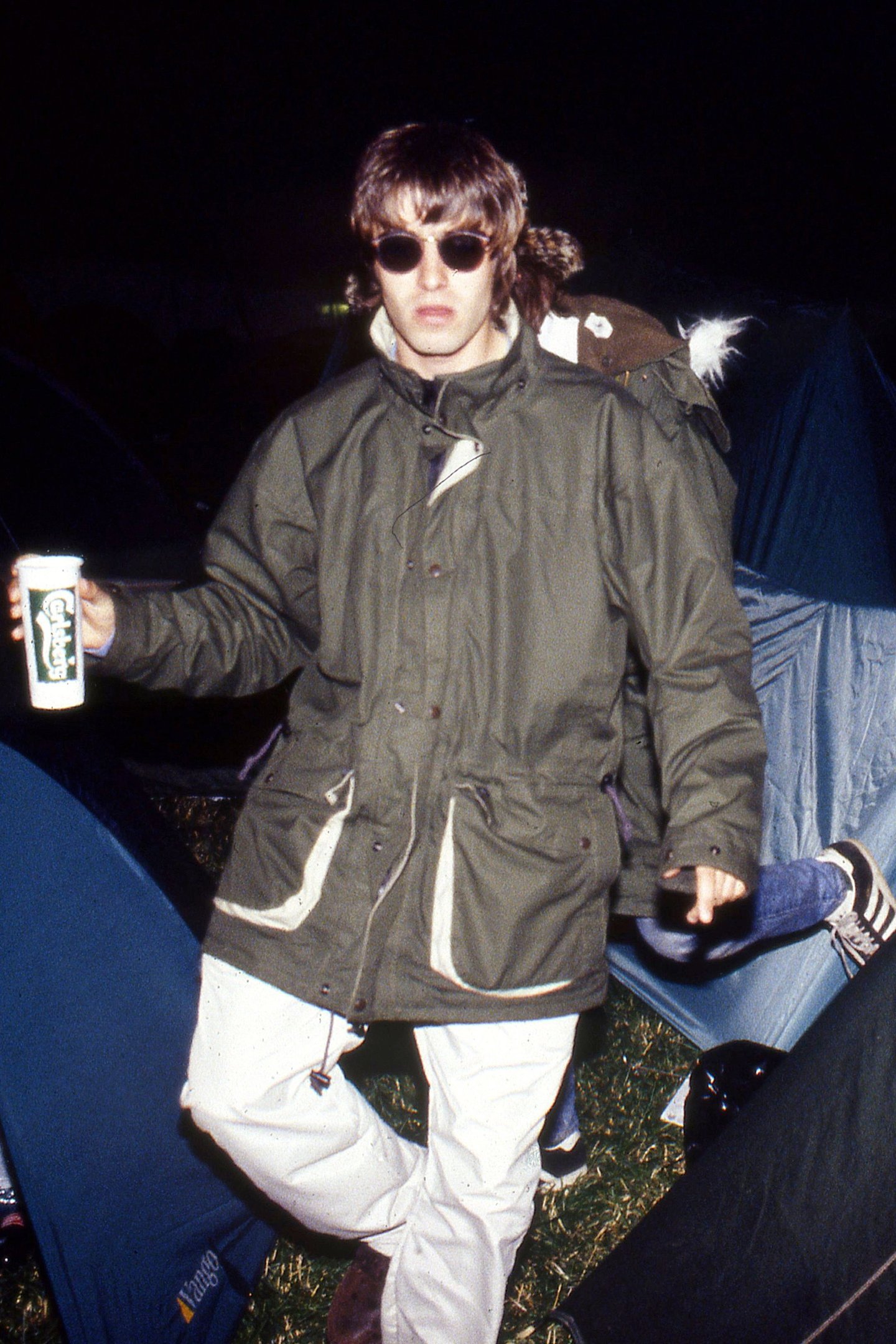 Jovem com óculos escuros e casaco verde segura lata de cerveja em acampamento, estilo britpop.