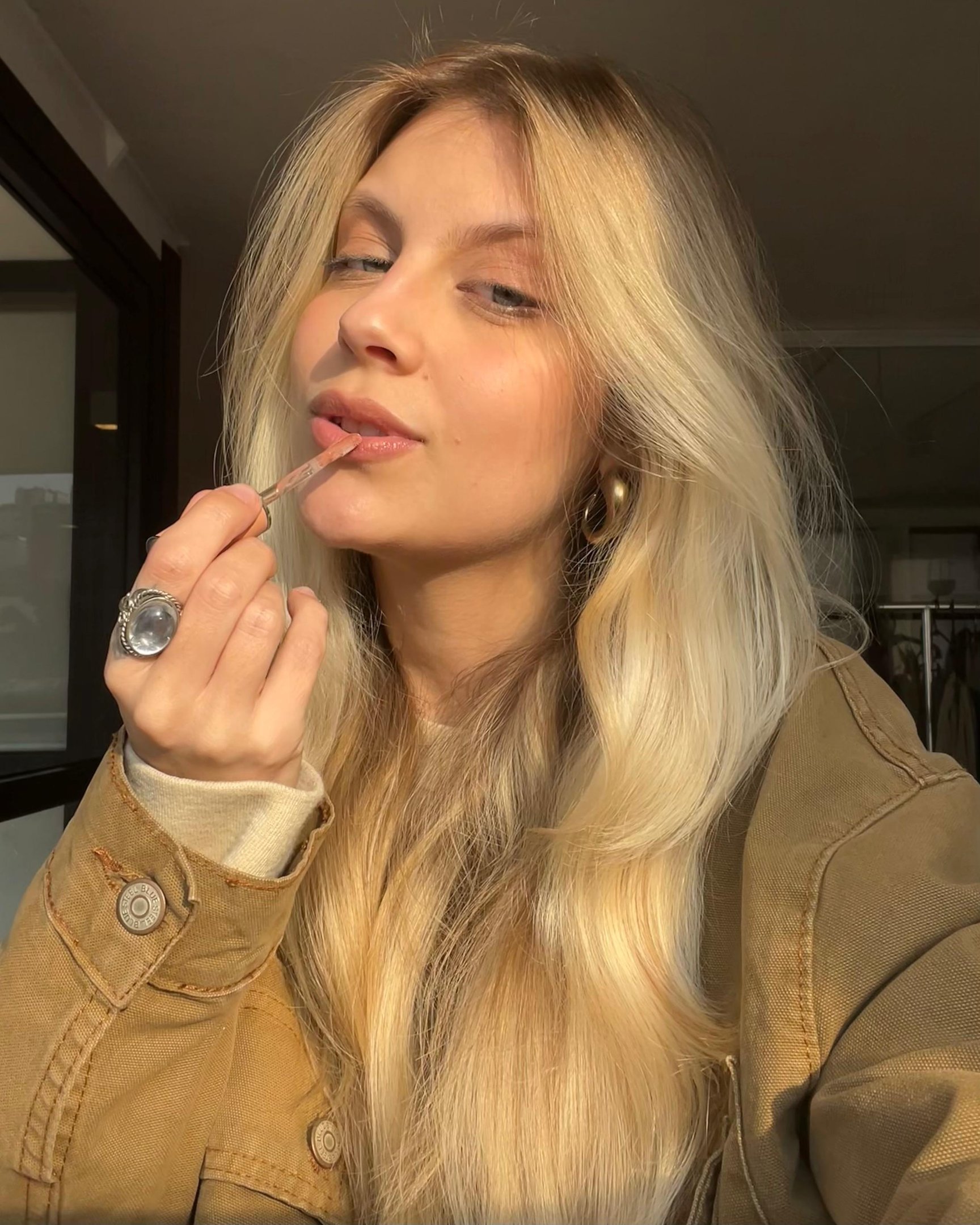 Mulher aplicando lip balm nos lábios, usando casaco marrom e anel prateado, sob luz natural suave.