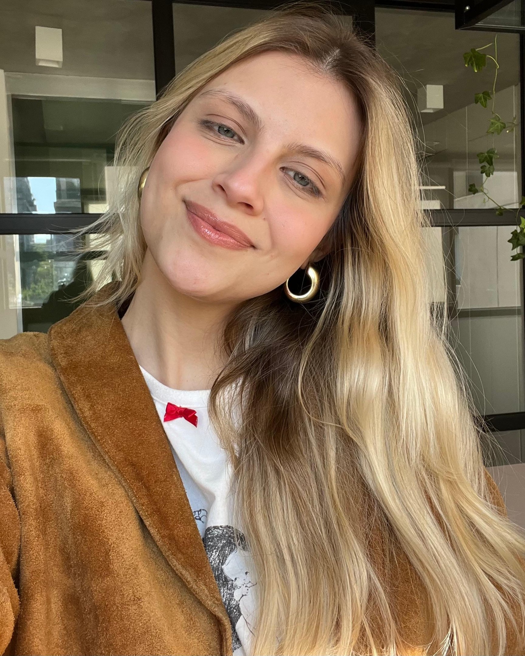 Moça sorrindo, lábios brilhantes com lip balm, veste casaco marrom e brincos de argola em ambiente interno.