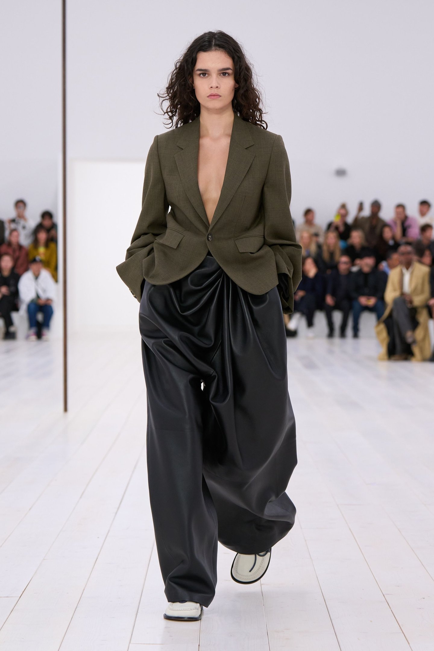 Modelo desfilando com blazer verde e calça balonê preta, em passarela branca cercada por público.