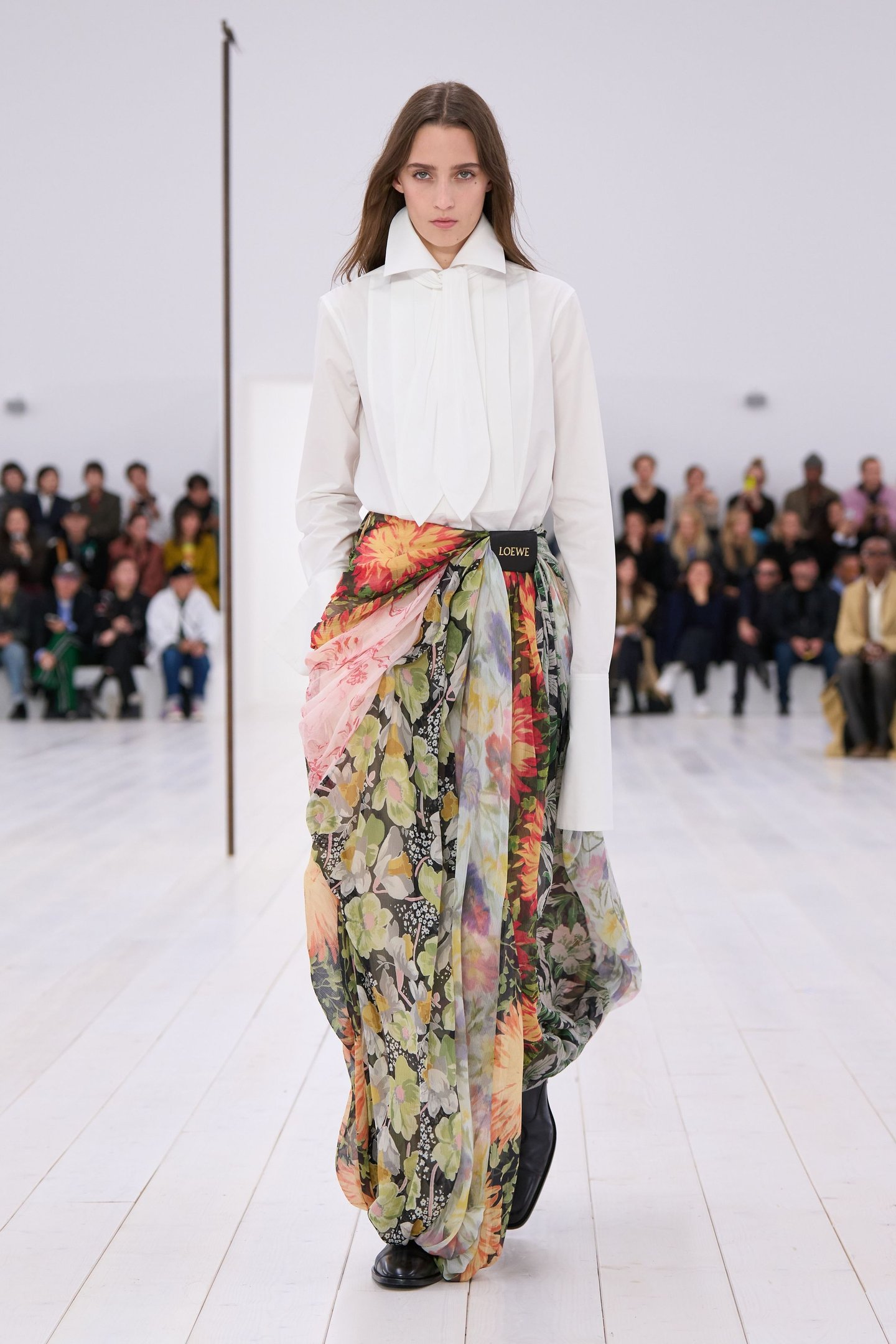 Modelo desfilando com blusa branca e saia longa floral transparente em passarela, diante de plateia.