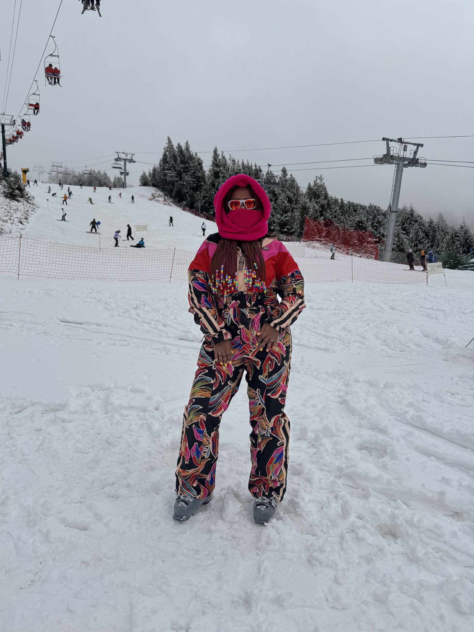 Pessoa em roupa de inverno colorida em pista de esqui, cercada por neve e teleféricos ao fundo. Looks de inverno coloridos.
