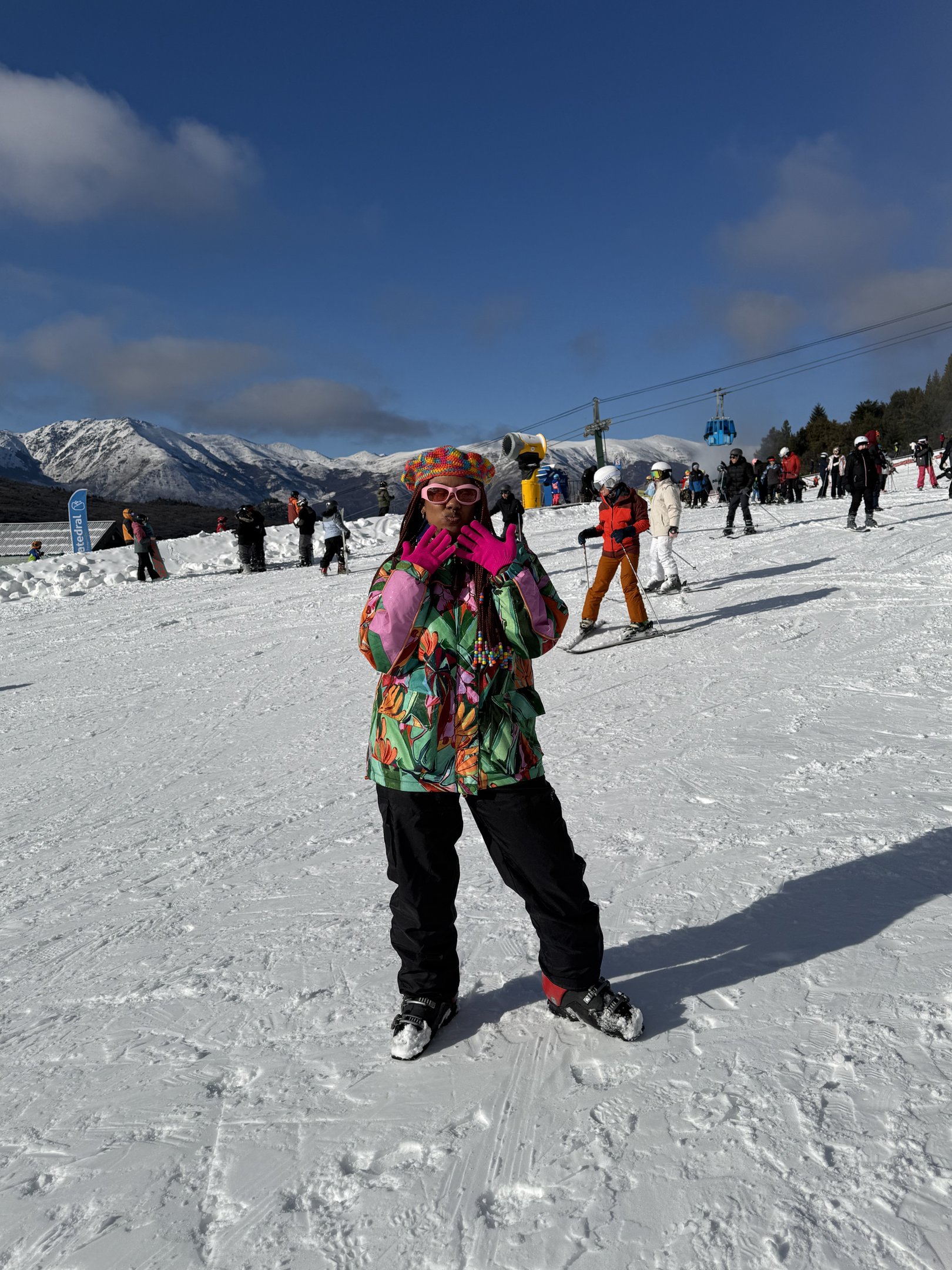 Pessoa em uma estação de esqui com roupa de inverno colorida, incluindo luvas rosa e jaqueta estampada. Looks de inverno coloridos