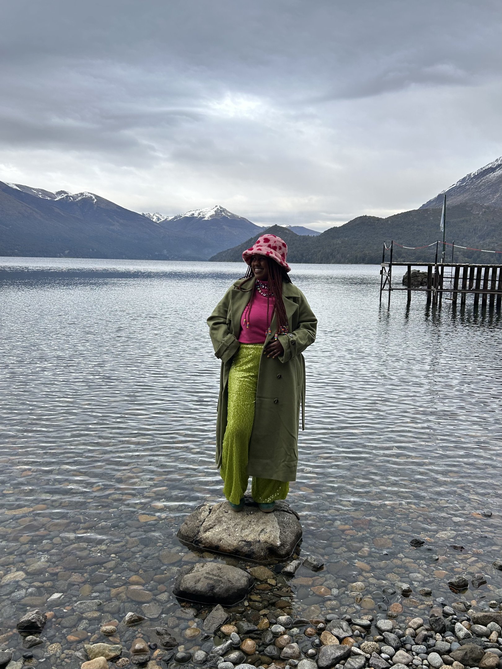 Pessoa usando casaco verde e calça verde em um lago, com montanhas ao fundo, estilo vibrante de inverno. Looks de inverno coloridos