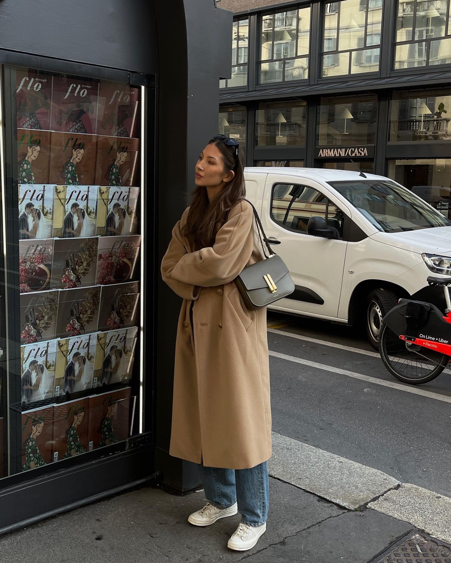 Pessoa com casaco bege e calça jeans ao lado de banca de revistas. Representa uma das ideias com looks com all star branco.