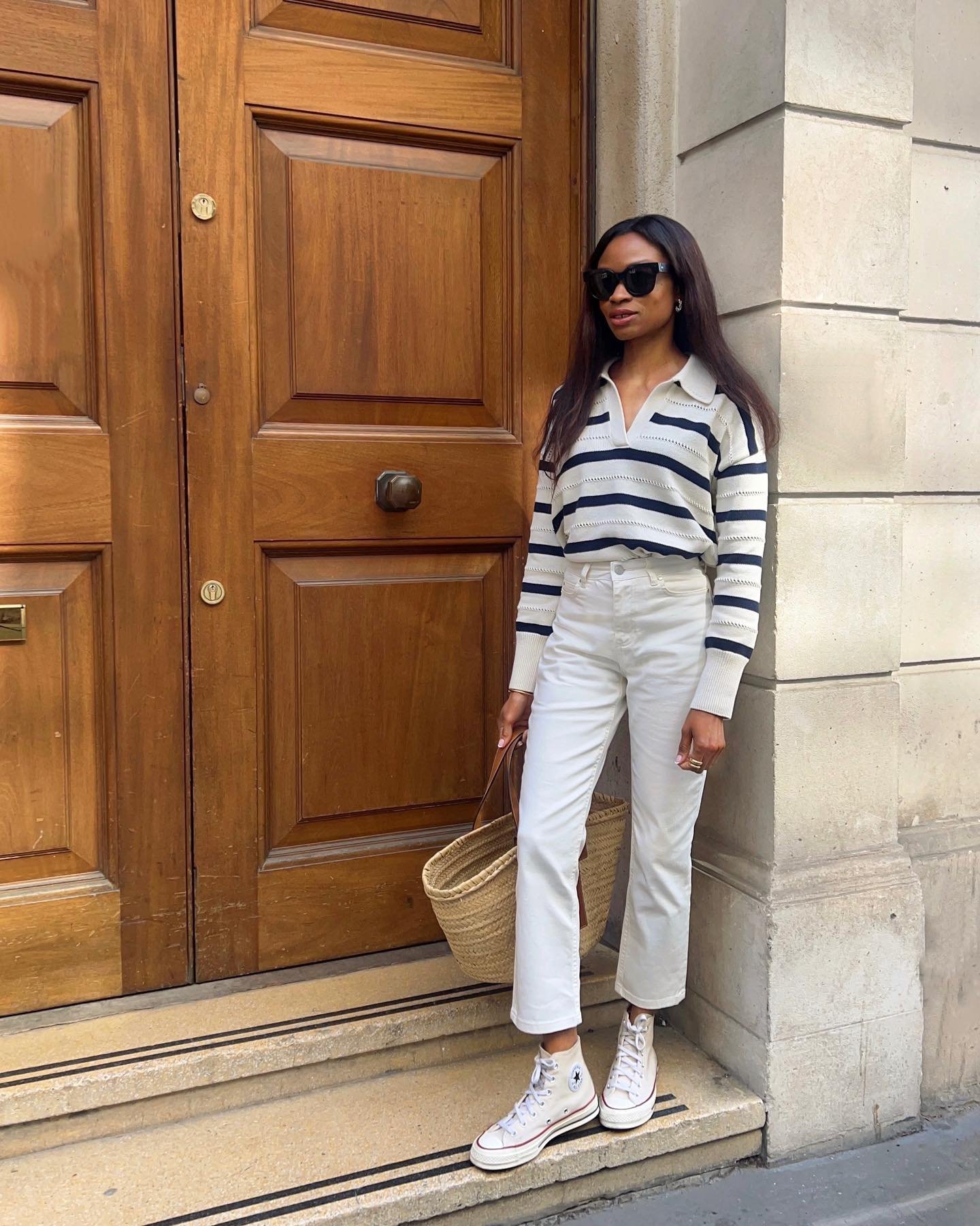 Pessoa com look casual, usando calça e blusa listrada, all star branco e bolsa de palha, em frente a uma porta de madeira.