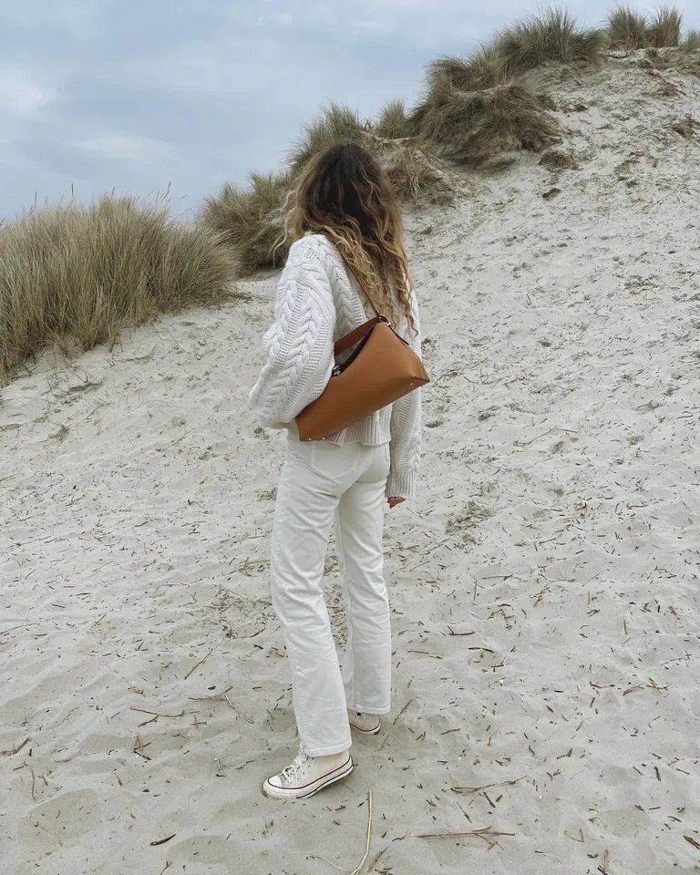 Mulher de cabelos longos com suéter e jeans brancos, usando All Star branco, em duna de areia.