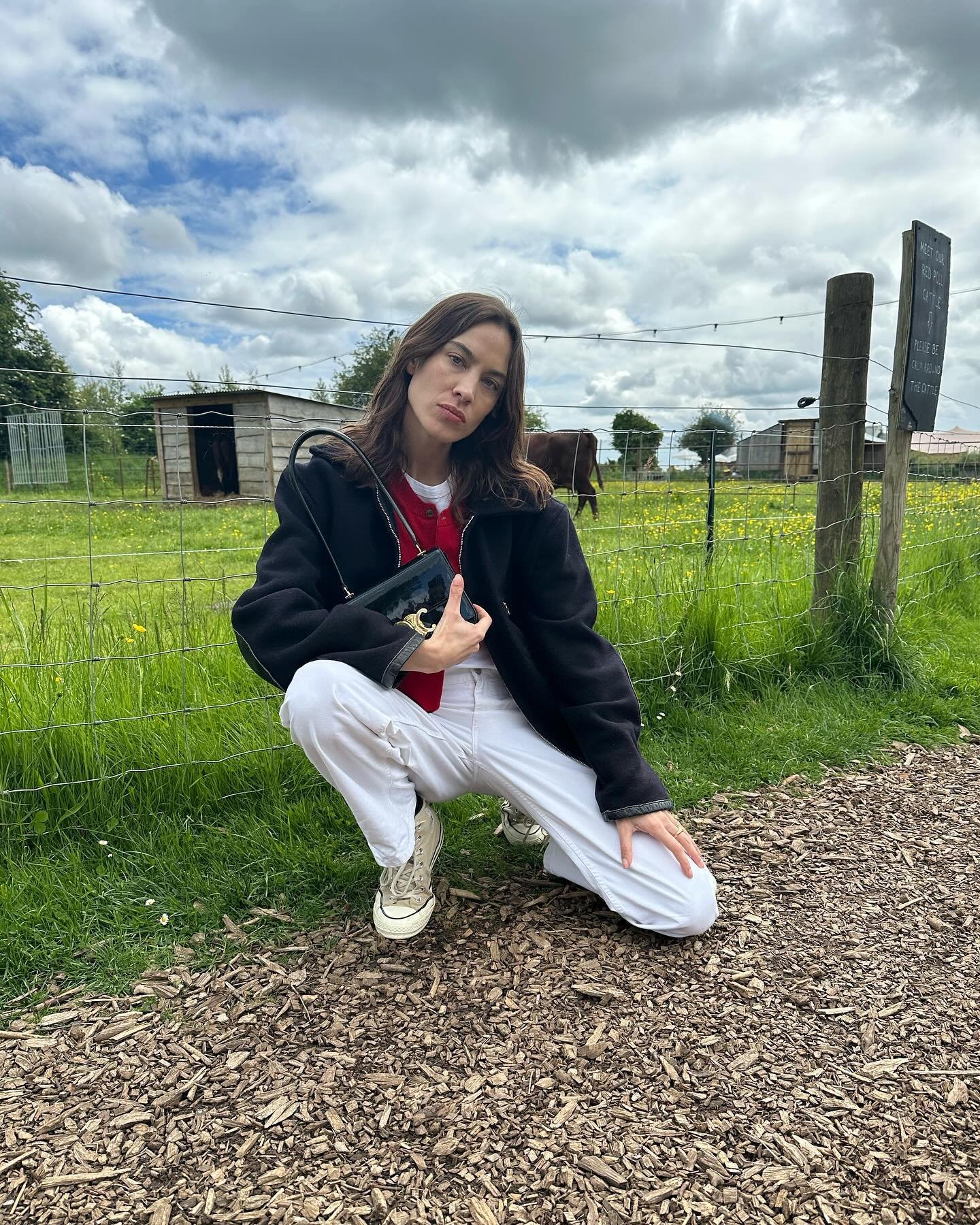 Pessoa agachada em campo com tênis branco, calça branca, casaco preto e bolsa, nuvens ao fundo.