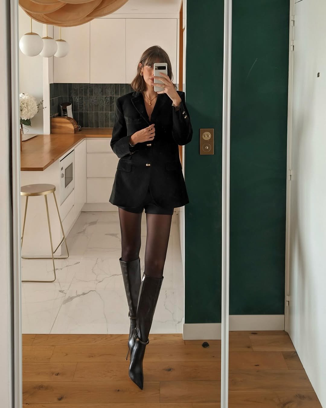 Pessoa usando blazer preto, shorts, meia-calça e botas de cano alto em frente a espelho.