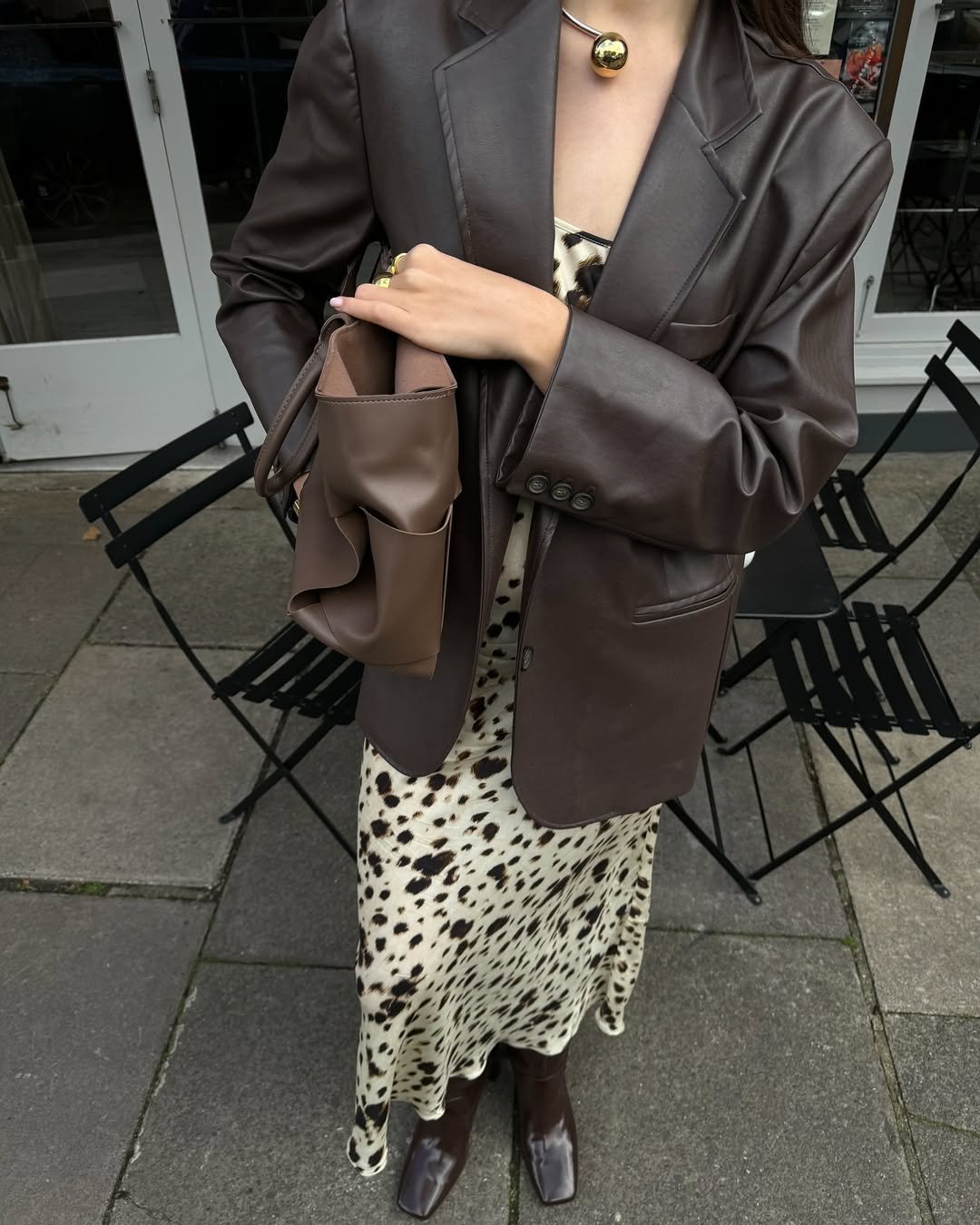 Pessoa com vestido estampado animal print, blazer marrom e botas de cano alto, segura bolsa marrom.