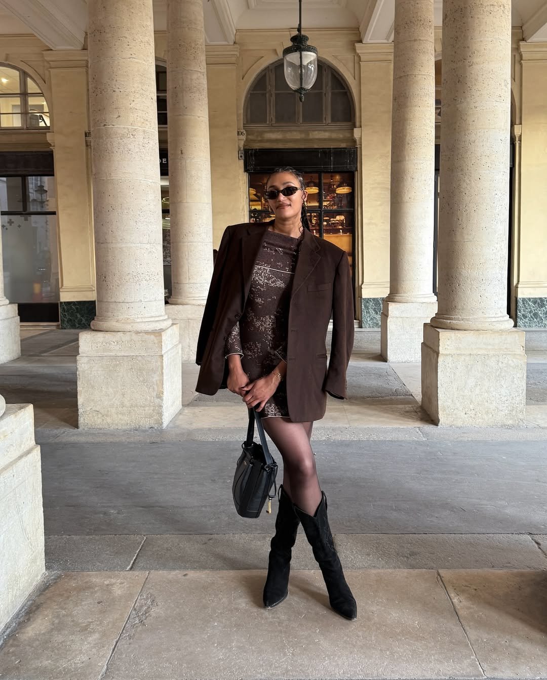 Pessoa em pé usando vestido marrom, blazer combinando, botas de cano alto e óculos escuros, demonstrando looks com bota cano alto.