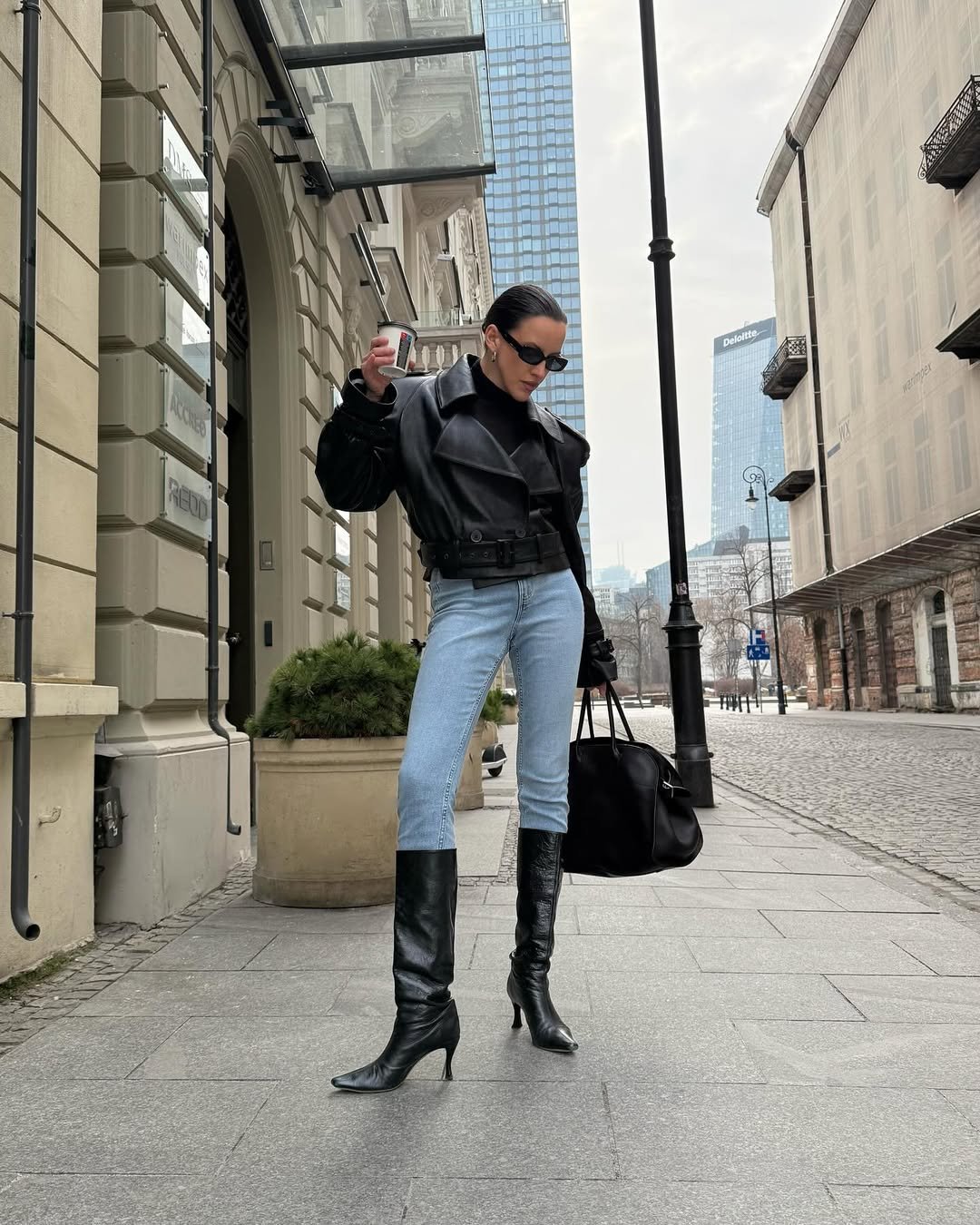 Pessoa com casaco preto, jeans e botas cano alto caminha em calçada urbana. Ela segura uma bebida.
