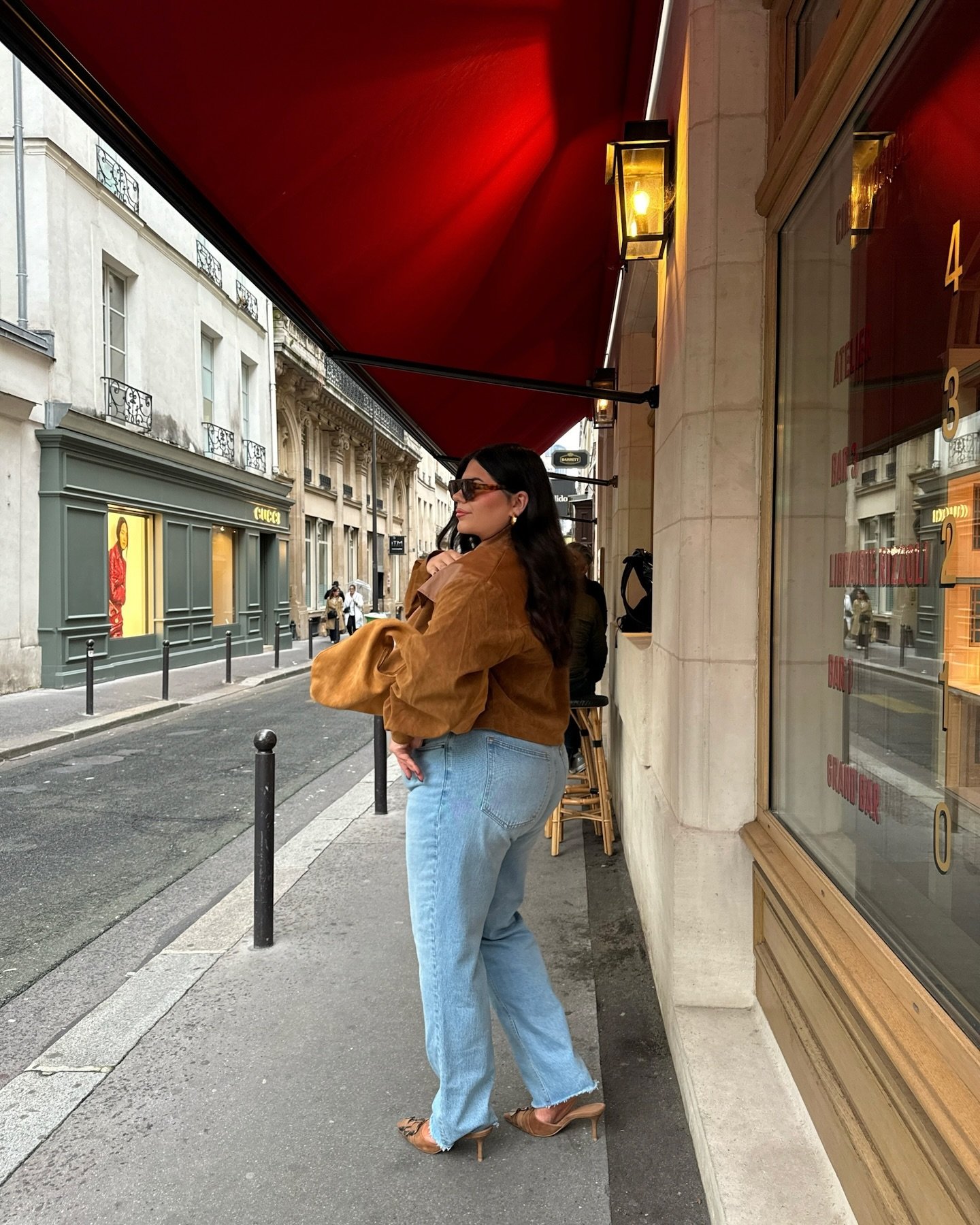 Pessoa usando blusa marrom e calça jeans, caminhando na calçada ao lado de uma loja.