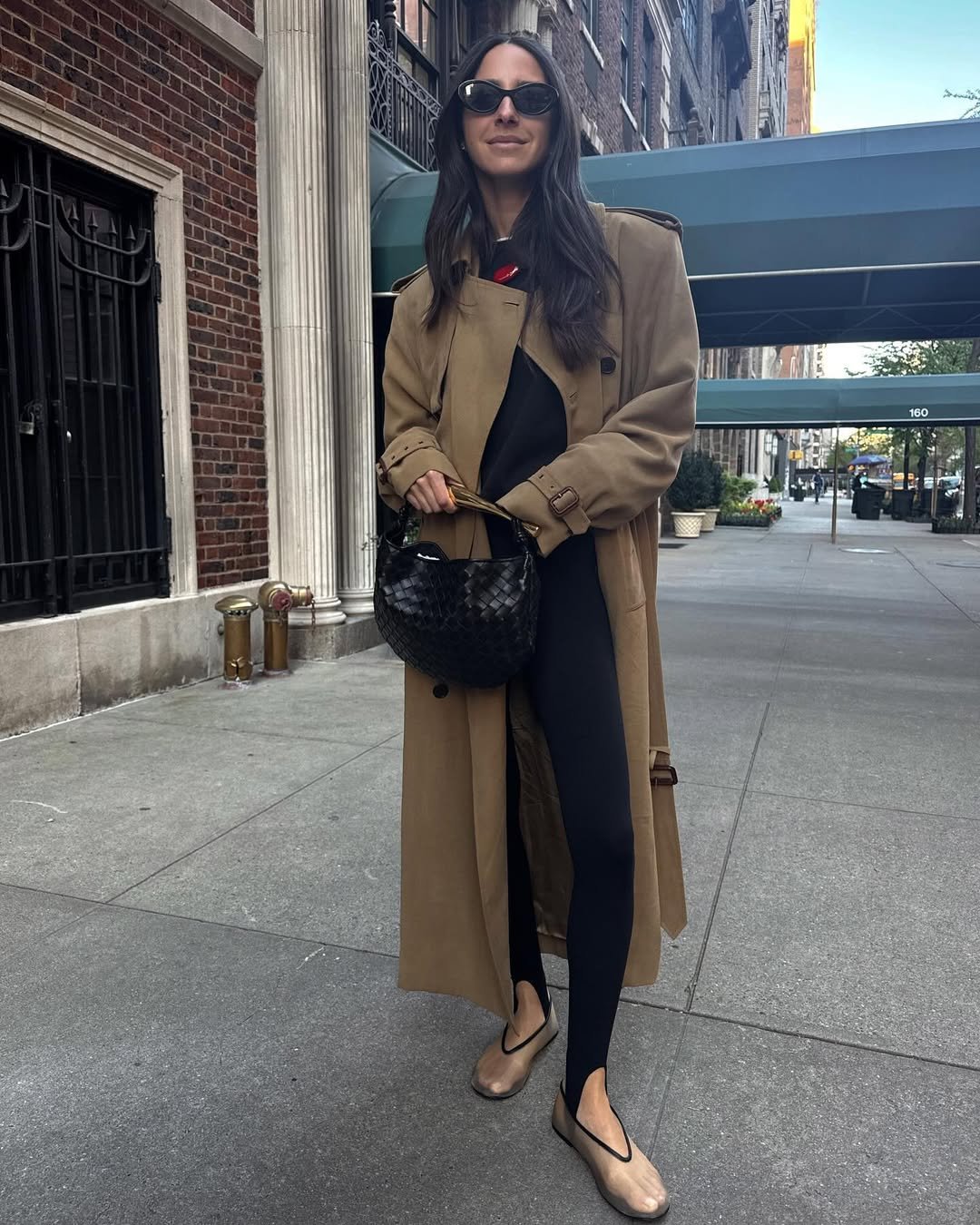 Mulher em look urbano, com casaco longo, calça legging preta e sapatos neutros, segura bolsa preta em rua da cidade.